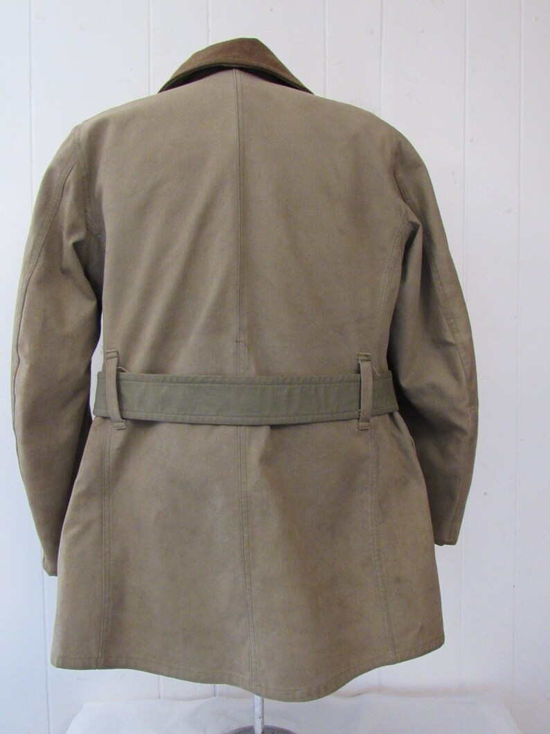 May include: A tan-colored jacket with a brown corduroy collar and a green canvas belt. The jacket has a long, straight cut and a single-breasted closure.