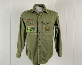 May include: Green long-sleeved button-up shirt with two patches. The left patch is a rectangular patch with the text "Wilderness Trail" and a scene of a forest. The right patch is a square patch with a gold star and the text "Scout".