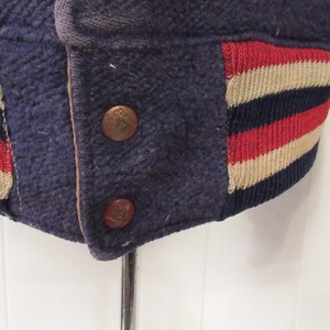 May include: Close-up of a navy blue varsity jacket with red, white, and navy blue striped ribbing. The jacket has two brass buttons.