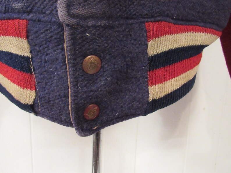 May include: Close-up of a navy blue varsity jacket with red, white, and navy blue striped ribbing. The jacket has two brass buttons.