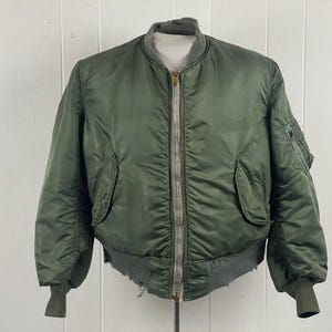 May include: Olive green bomber jacket with a ribbed collar, cuffs, and waistband. The jacket features a full-length zipper, two front pockets, and a small pocket on the left sleeve. The jacket is made of a smooth, possibly nylon, material.