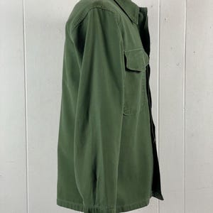 May include: Olive green button-down shirt with a collared neckline and a single chest pocket. The long-sleeved shirt is made of a durable fabric and has a classic, utilitarian design. The shirt is displayed on a mannequin against a white background.