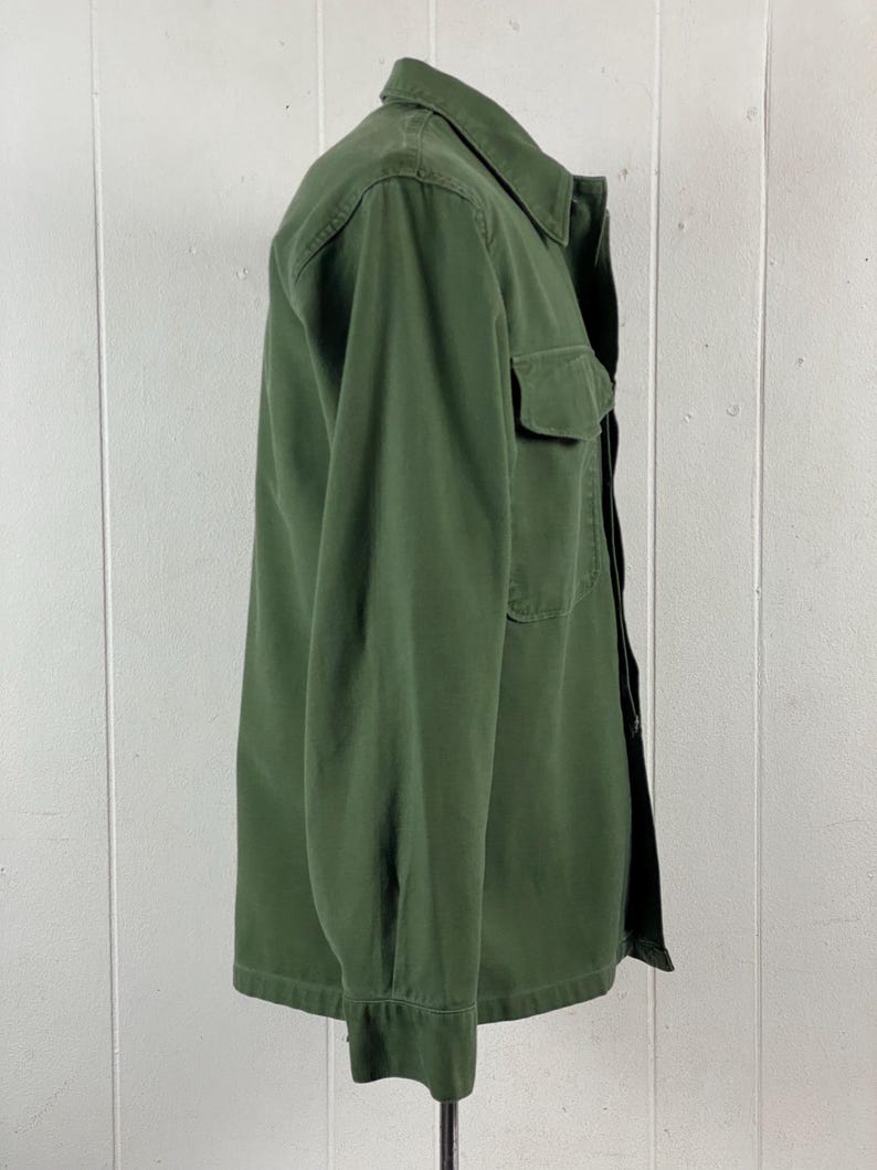 May include: Olive green button-down shirt with a collared neckline and a single chest pocket. The long-sleeved shirt is made of a durable fabric and has a classic, utilitarian design. The shirt is displayed on a mannequin against a white background.