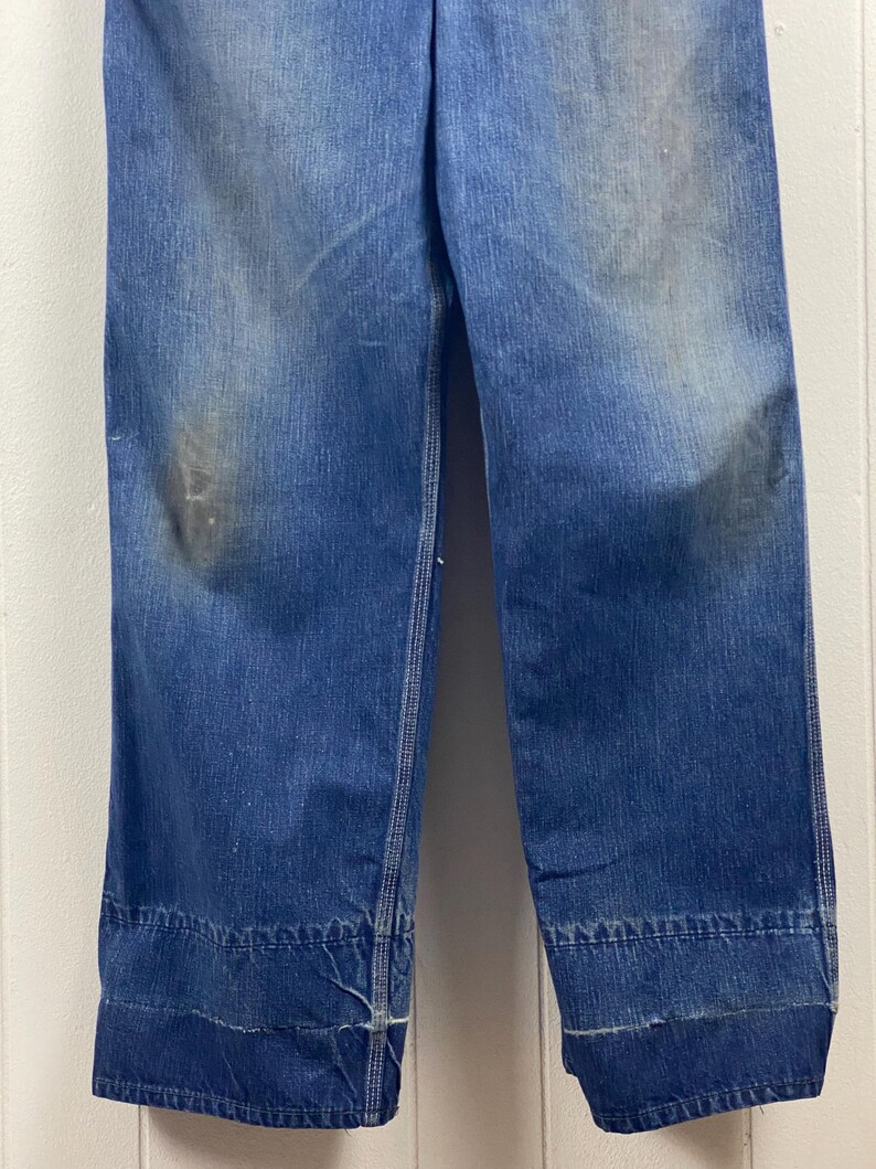 K&ouml;nnte beinhalten: Nahaufnahme einer Vintage-Jeans aus blauem Denim. Die Jeans hat eine verblasste Waschung mit Gebrauchsspuren und ein weites Bein. Die S&auml;ume sind umgeschlagen und mit wei&szlig;em Faden gen&auml;ht. Der Denim hat einen klassischen, strapazierf&auml;higen Look.