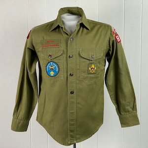 May include: Green Boy Scout uniform shirt with long sleeves and a button-up front. The shirt has several patches, including a "SERVE" patch, a "Campfire" patch, and a Scout emblem patch.