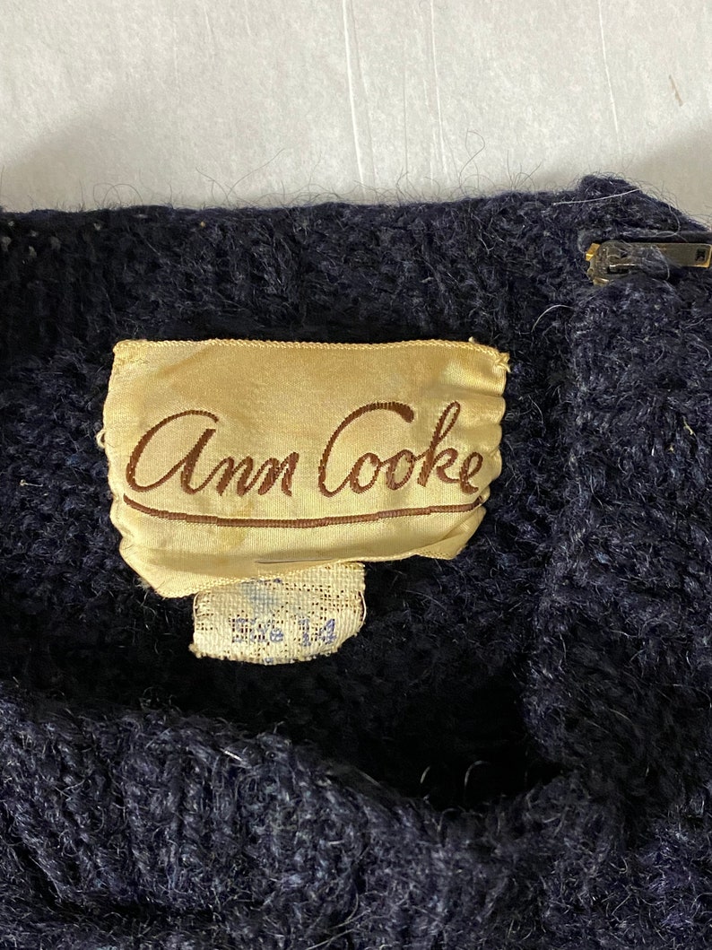 May include: A close-up of a dark blue knit sweater with a tan label that reads 'Ann Cooke' in brown cursive lettering. The label also has a smaller white tag that reads 'Size 14'.