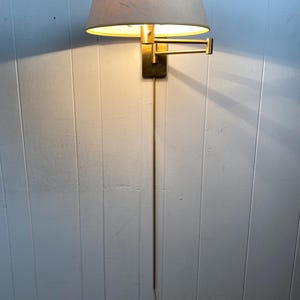 May include: A vintage wall lamp with a brass finish and a beige lampshade. The lamp has an adjustable arm and a long cord. The light is on, casting a warm glow. The lamp is mounted on a white panelled wall.