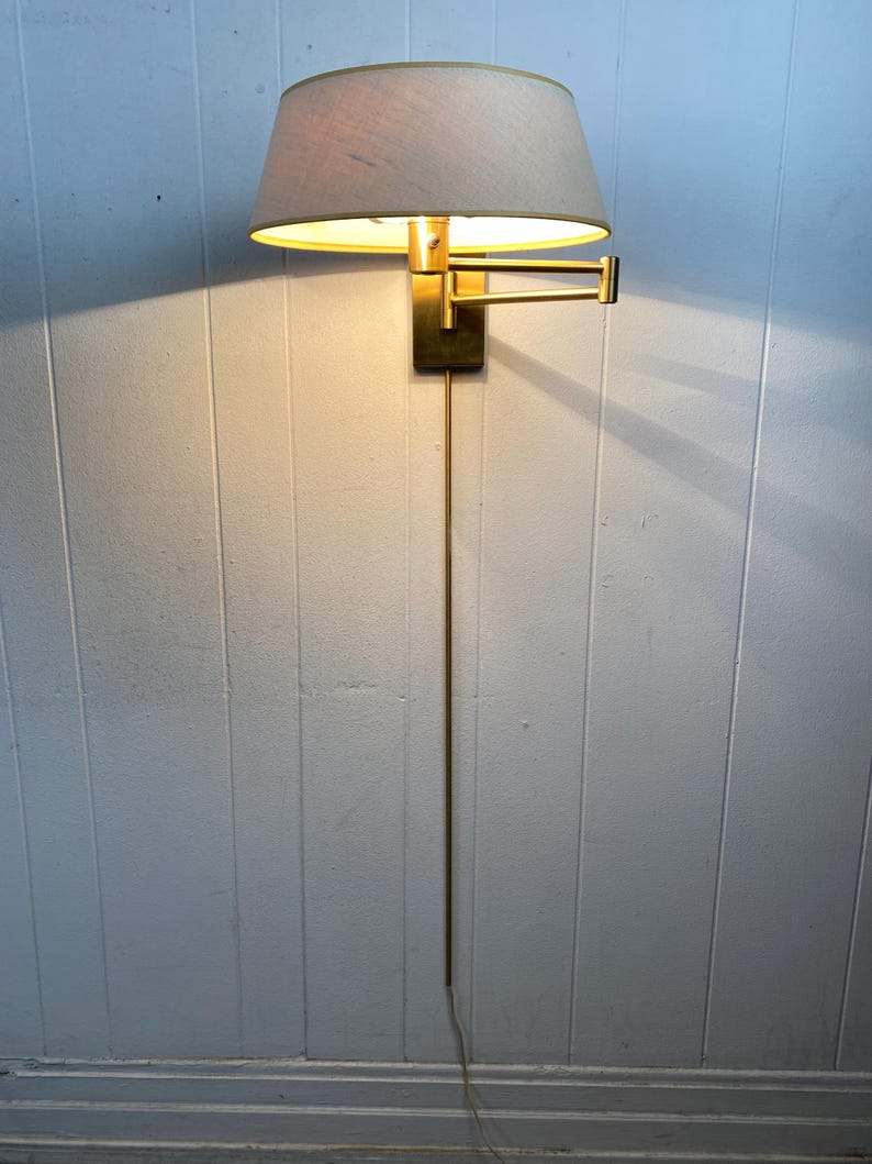 May include: A vintage wall lamp with a brass finish and a beige lampshade. The lamp has an adjustable arm and a long cord. The light is on, casting a warm glow. The lamp is mounted on a white panelled wall.