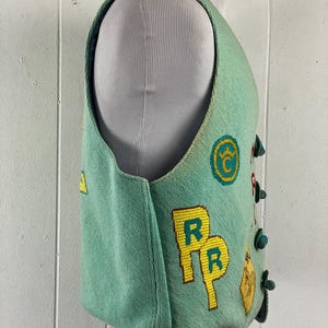 May include: A teal blue vest with yellow and green embroidered patches. The patches include letters, symbols, and a railroad crossing sign.