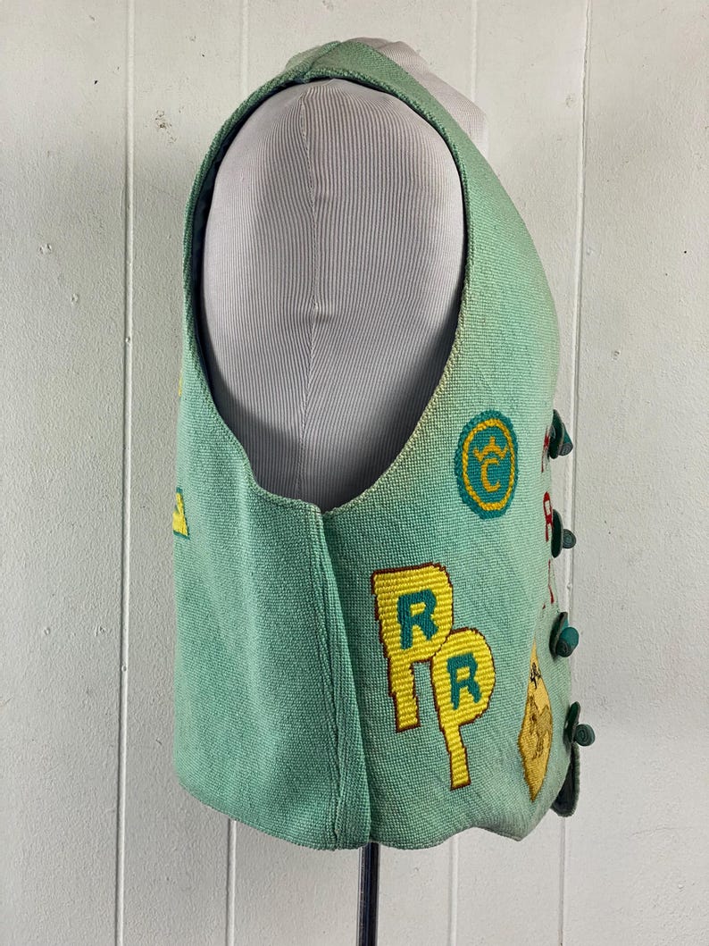 May include: A teal blue vest with yellow and green embroidered patches. The patches include letters, symbols, and a railroad crossing sign.