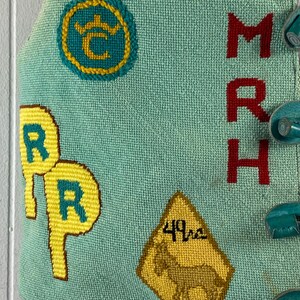 May include: A turquoise blue fabric with embroidered details. The fabric features a yellow circle with a crown and the letter "C" inside, two yellow circles with the letter "R" inside, a yellow diamond-shaped sign with the text "49ers" and a brown horse inside, and the letters "MRH" in red.