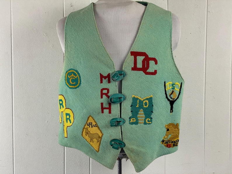 May include: A green vest with embroidered patches and buttons. The patches include letters, a crown, a horse, and a road sign. The vest has a V-neck and is made of a soft, textured fabric.