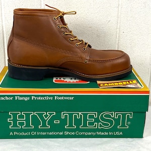 May include: A brown leather work boot with yellow laces, sitting on a green and yellow box. The box reads "HY-TEST" and "Anchor Flange Protective Footwear". The boot has a black sole and a padded collar.