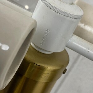 May include: Close-up of a white and brass lamp fixture. The white components include a cylindrical socket with the text "Nessen" and a curved ceramic piece. The brass base is cylindrical.