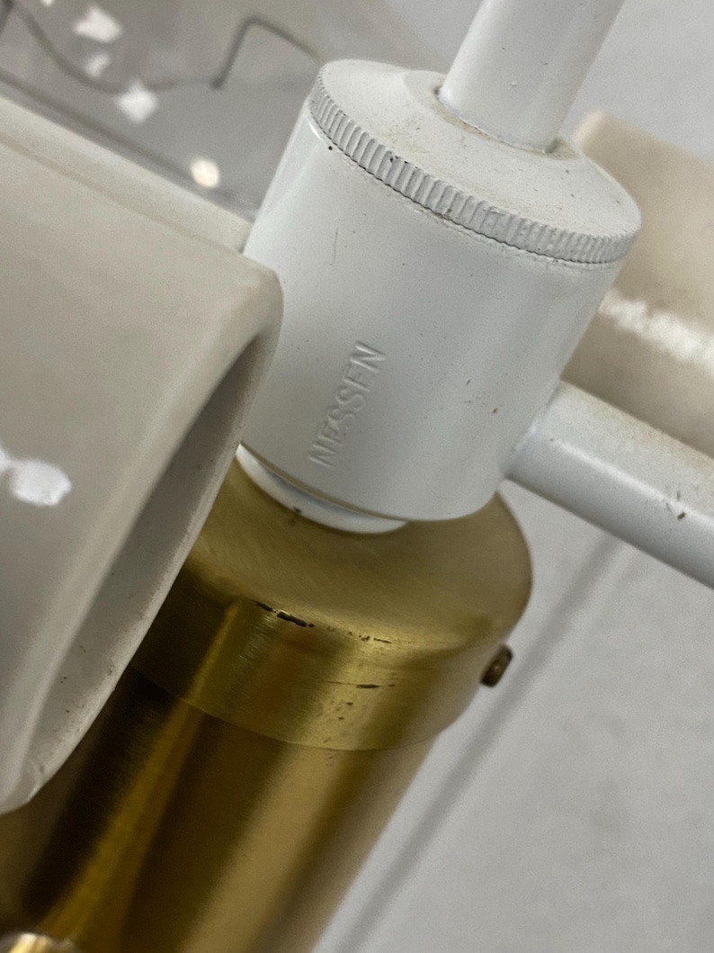 May include: Close-up of a white and brass lamp fixture. The white components include a cylindrical socket with the text "Nessen" and a curved ceramic piece. The brass base is cylindrical.