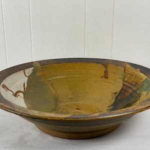 Op de afbeelding: Een grote, bruine en witte keramische kom met een brede rand en een licht uitlopende lip. De kom heeft een rustieke, handgeworpen uitstraling en is geglazuurd in tinten bruin, wit en groen.