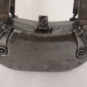 May include: A silver-colored,  half-moon shaped clutch purse with a decorative clasp and ornate silver handles. The purse has a marbled, textured surface.