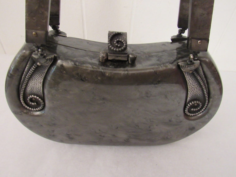 May include: A silver-colored,  half-moon shaped clutch purse with a decorative clasp and ornate silver handles. The purse has a marbled, textured surface.