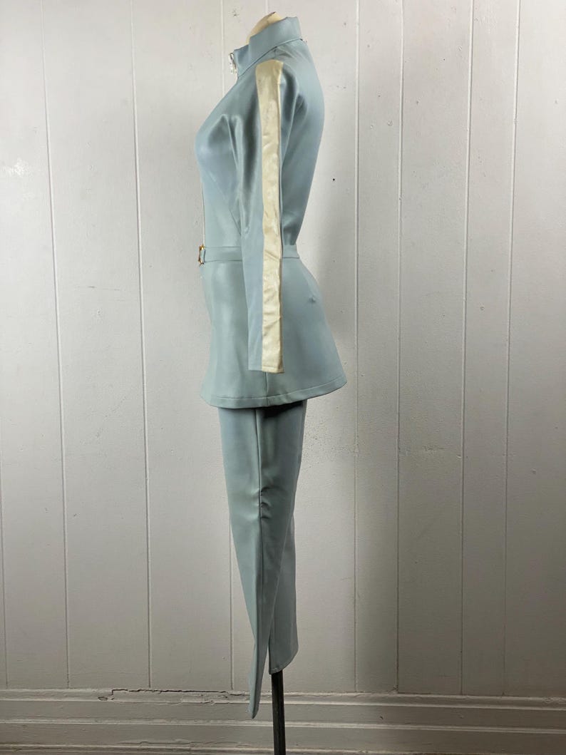 K&ouml;nnte beinhalten: Ein hellblauer Jumpsuit mit einem wei&szlig;en Streifen an der Seite jedes &Auml;rmels. Der Jumpsuit hat einen Rei&szlig;verschluss und einen G&uuml;rtel.