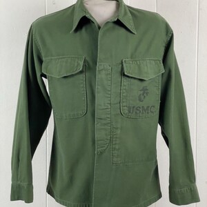 May include: Olive green military-style shirt with two chest pockets and a collared neckline. The shirt features a USMC emblem and the letters "USMC" printed on the right chest pocket. The long-sleeved shirt is button-down.