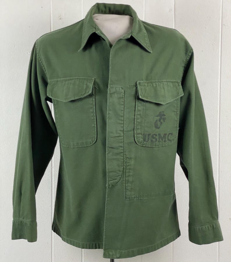 May include: Olive green military-style shirt with two chest pockets and a collared neckline. The shirt features a USMC emblem and the letters "USMC" printed on the right chest pocket. The long-sleeved shirt is button-down.