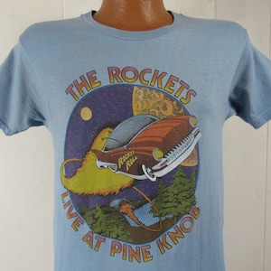 May include: A light blue t-shirt featuring a graphic of a car flying through space with the text "The Rockets Live At Pine Knob".