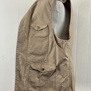 May include: A tan-colored vest with multiple pockets and button closures. The vest is made of a lightweight fabric and has a classic design. It is displayed on a mannequin, showcasing its shape and fit. The vest appears to be in good condition.