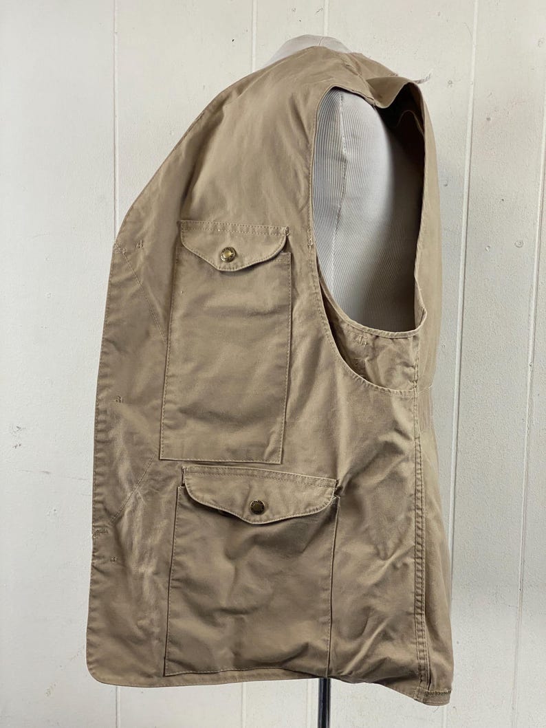 May include: A tan-colored vest with multiple pockets and button closures. The vest is made of a lightweight fabric and has a classic design. It is displayed on a mannequin, showcasing its shape and fit. The vest appears to be in good condition.