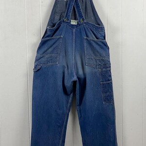 K&ouml;nnte beinhalten: Vintage blaue Jeans-Latzhose mit verstellbaren Tr&auml;gern. Die Latzhose hat mehrere Taschen, darunter eine Latz- und Seitentaschen. Die Tr&auml;ger tragen das Logo "OshKosh B'gosh". Die Manschetten haben einen verblichenen wei&szlig;en Streifen.