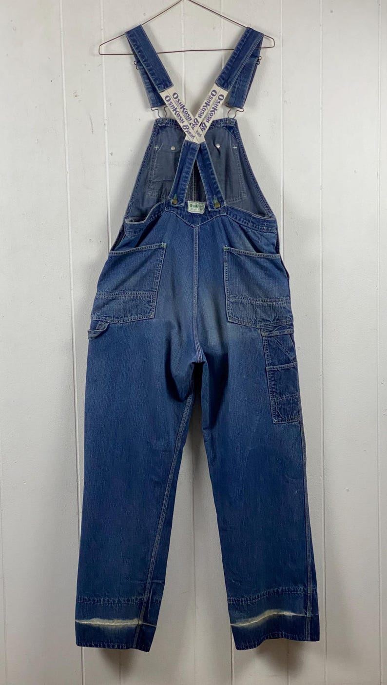 K&ouml;nnte beinhalten: Vintage blaue Jeans-Latzhose mit verstellbaren Tr&auml;gern. Die Latzhose hat mehrere Taschen, darunter eine Latz- und Seitentaschen. Die Tr&auml;ger tragen das Logo "OshKosh B'gosh". Die Manschetten haben einen verblichenen wei&szlig;en Streifen.
