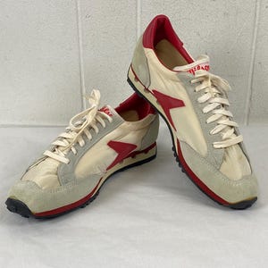 May include: Pair of vintage athletic shoes. The sneakers feature a cream-colored nylon upper with red accents and a red lightning bolt design. The toe and heel are reinforced with gray suede, and the soles are black and red.