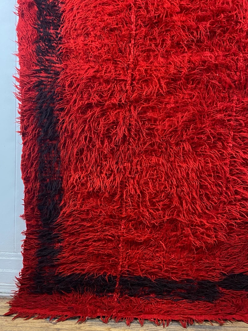 May include: A red shag rug with a black border. The rug has a long, fluffy pile and is made of wool.