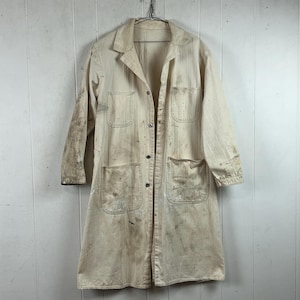 May include: A long, off-white work coat featuring a collared neckline, long sleeves, and a front button closure. The garment displays multiple patch pockets on the chest and hips, with visible paint splatters and stains across the fabric surface.
