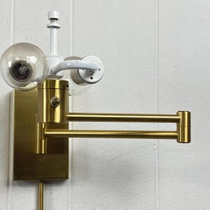 May include: A brass-toned wall lamp with a pivoting arm and a clear globe bulb. The lamp has a rectangular base and a white arm holding a second bulb. The arm extends approximately 30 cm.