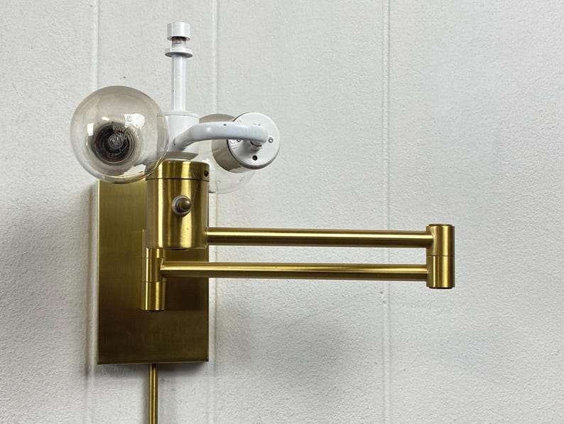 May include: A brass-toned wall lamp with a pivoting arm and a clear globe bulb. The lamp has a rectangular base and a white arm holding a second bulb. The arm extends approximately 30 cm.