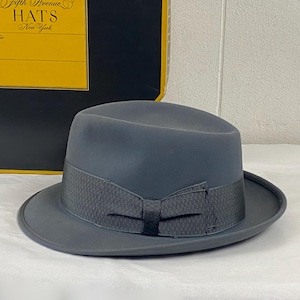 Op de afbeelding: Een grijze fedora hoed met een donkergrijze lintband en strik. De hoed wordt gepresenteerd voor een zwart-gele doos met de tekst "Fifth Avenue Hats New York".