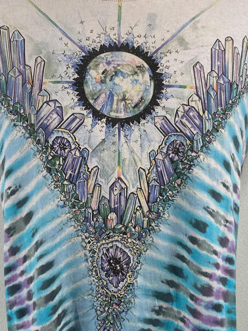 Puede incluir: Una camiseta tie-dye con un dise&ntilde;o de cristales coloridos y una imagen circular de un planeta en el centro.