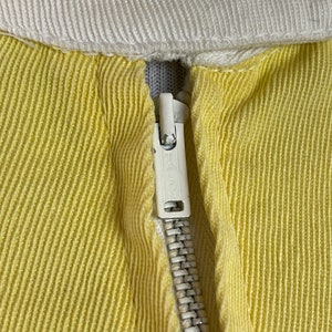 May include: Close-up of a yellow zipper on a garment. The zipper is metal with a white pull tab. The fabric is a yellow ribbed material.