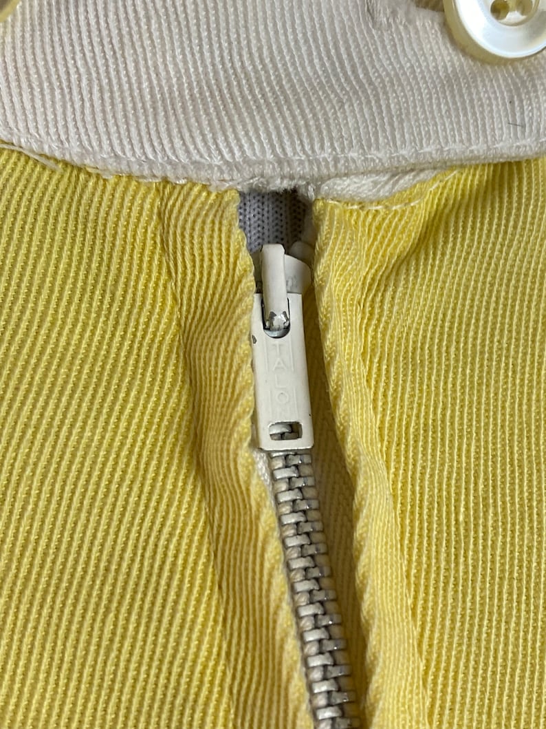 May include: Close-up of a yellow zipper on a garment. The zipper is metal with a white pull tab. The fabric is a yellow ribbed material.