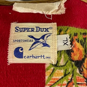 以下が含まれることがあります： 赤と緑の模様の生地のクローズアップ。Super Dux Sportswearのラベルが付いています。ラベルには青いアヒルのグラフィックとCarhartt, Inc.のロゴが入っています。白いタグにはXLサイズと記載されています。