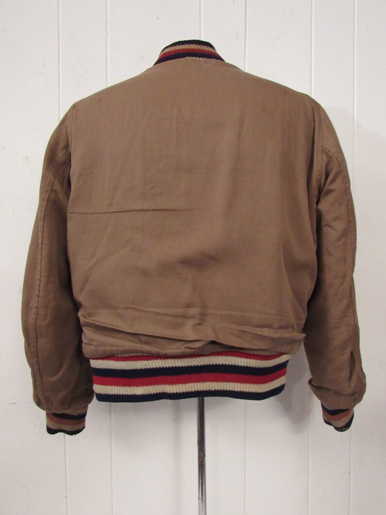 May include: A brown bomber jacket with a ribbed collar and cuffs. The cuffs have a navy, white, and red stripe pattern.