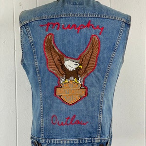 Può includere: Gilet di jeans blu con ricamo rosso che dice "Murphy Outlaw" e una toppa di aquila Harley Davidson.