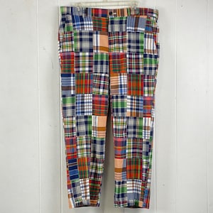 以下が含まれることがあります： 青、緑、赤、オレンジ、白の色合いのチェック柄が様々なパッチワークパンツ。白いハンガーにかけられ、白い背景に吊るされています。
