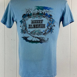 K&ouml;nnte beinhalten: Hellblaues T-Shirt mit einem grafischen Aufdruck einer Bergszene und dem Text "Banff Alberta".