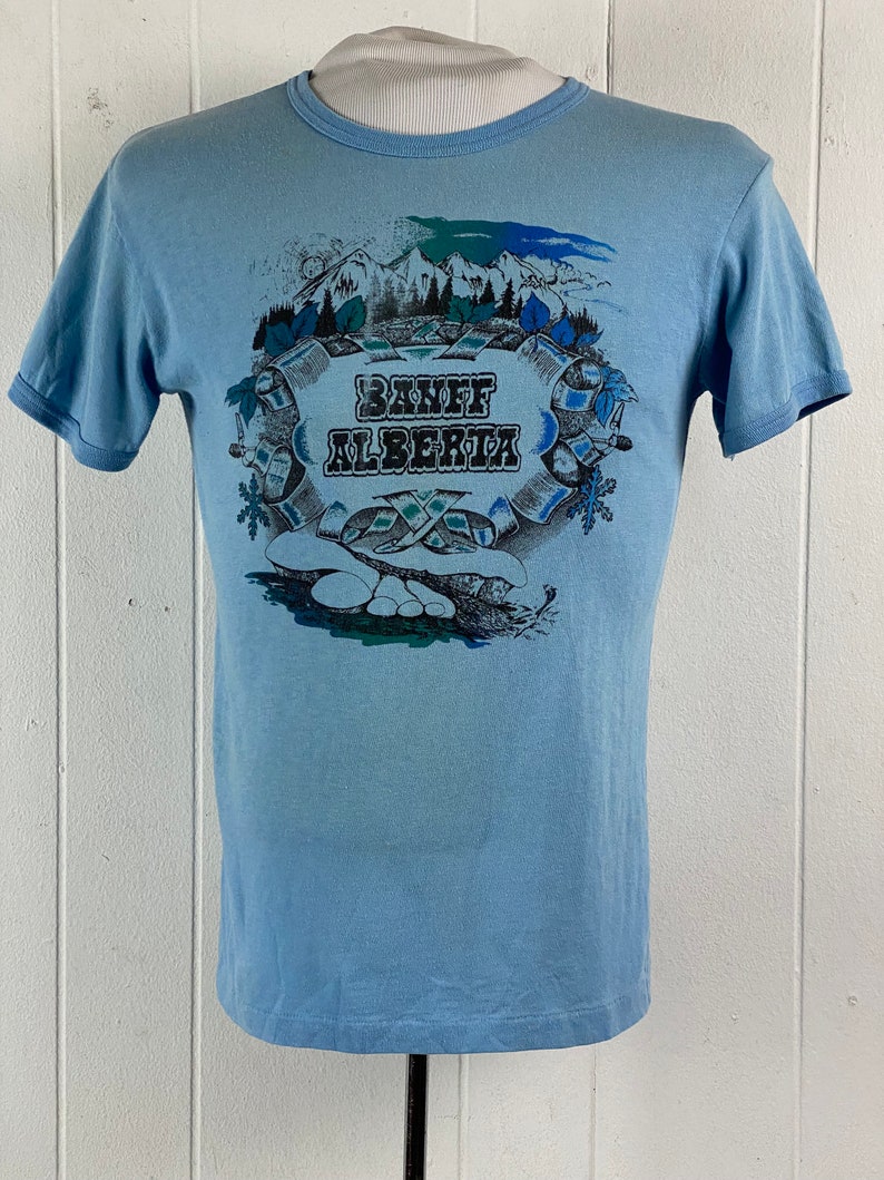 K&ouml;nnte beinhalten: Hellblaues T-Shirt mit einem grafischen Aufdruck einer Bergszene und dem Text "Banff Alberta".