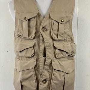 May include: A tan-colored vest with multiple pockets and button closures. The vest has a V-neck design and is displayed on a mannequin. The pockets are of varying sizes and are secured with flaps and buttons. The vest appears to be made of a durable, possibly canvas-like material.