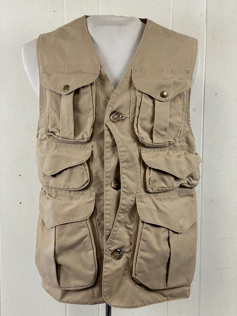 May include: A tan-colored vest with multiple pockets and button closures. The vest has a V-neck design and is displayed on a mannequin. The pockets are of varying sizes and are secured with flaps and buttons. The vest appears to be made of a durable, possibly canvas-like material.