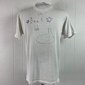 Könnte beinhalten: Weißes T-Shirt mit dem Text "S.C. Beat It" und einem lila Stern. Auf dem Shirt ist ein handgezeichnetes Bild einer Hand.