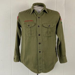 May include: Olive green button-down shirt with two chest pockets and long sleeves. The shirt has a collar and a patch on the left chest that reads "Boy Scouts of America" in red lettering. Patches are also visible on the sleeves.