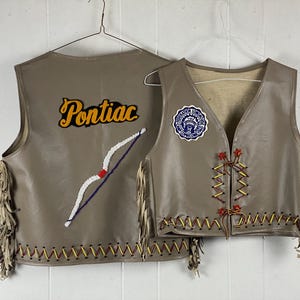 Peut inclure: Deux gilets en cuir beige avec franges et coutures décoratives. L'un des gilets porte le mot "Pontiac" en écriture jaune et un arc et des flèches perlés. L'autre gilet a un écusson circulaire avec du texte.